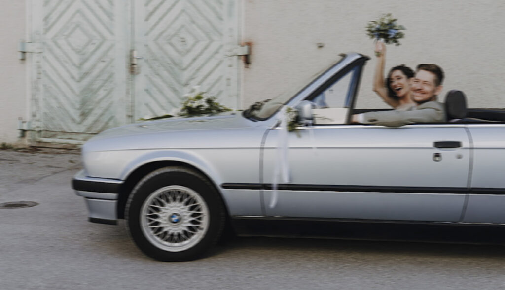 Das Hochzeitspaar im Brautauto. Ein alter BMW in silber geschmückt mit Blumen. Das Brautpaar lacht in die Kamera und die Braut streckt den Brautstrauß in die Höhe.